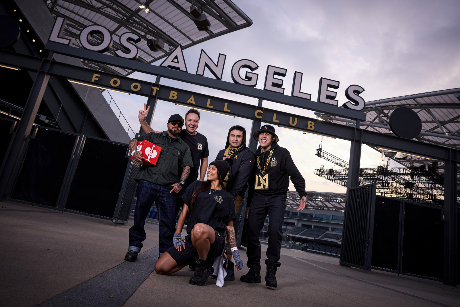 Five enthusiastic LAFC fans pose in front of the Los Angeles Football Club stadium entrance, wearing team gear and smiling, with the stadiums large sign visible above them.