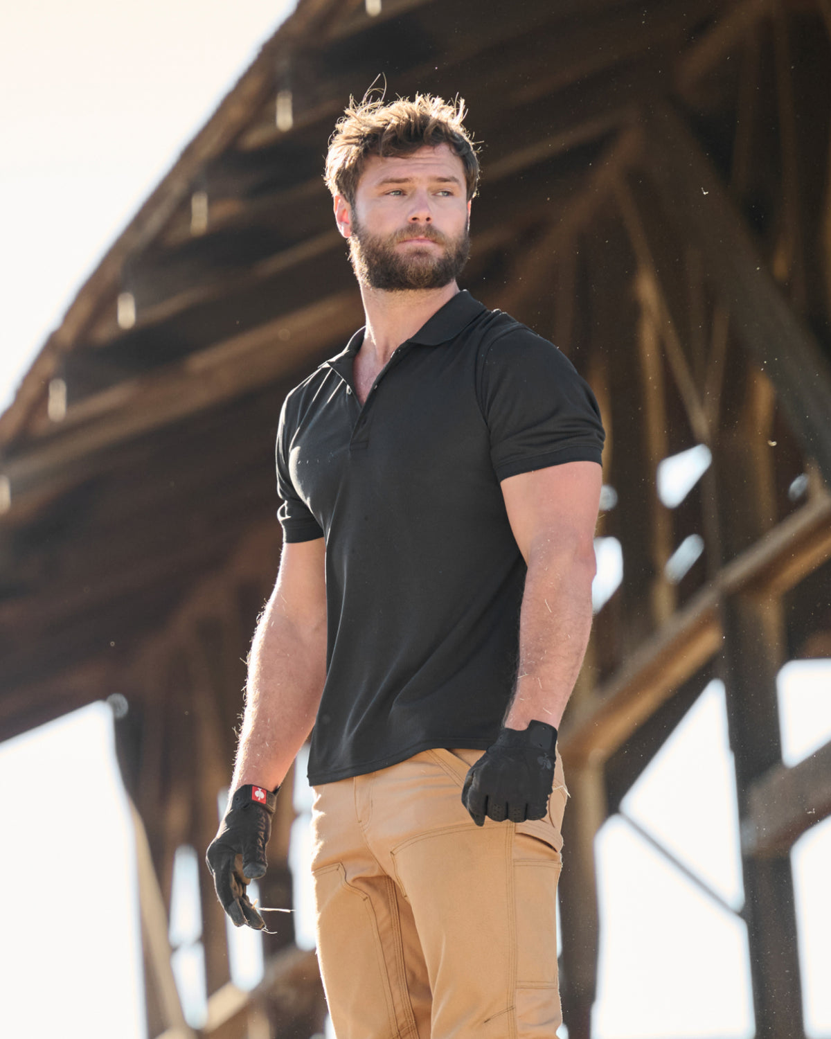A worker wears the STRAUSS Performance Polo Shirt in black with tan work pants on a sunny jobsite