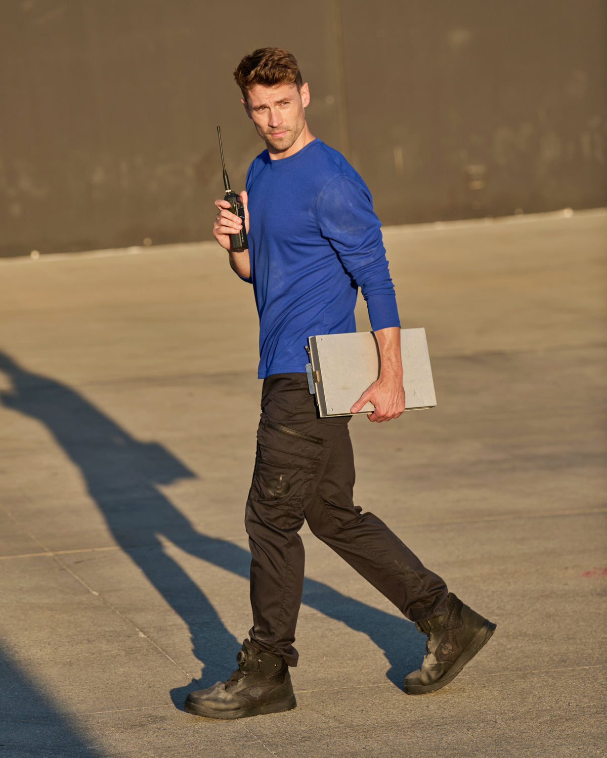 A worker wears the STRAUSS Performance Long Sleeve Shirt in royal blue with tan work pants beside jobsite equipment