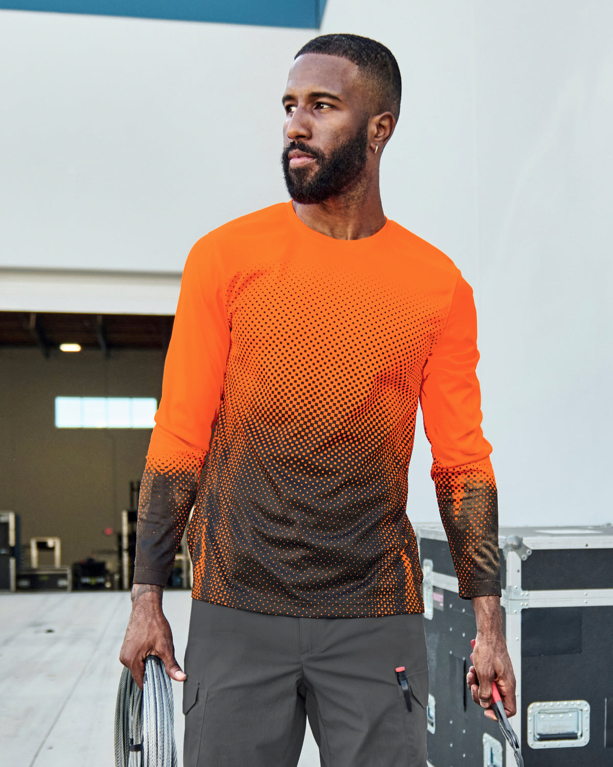 A worker wears the STRAUSS Enhanced Visibility Performance Long Sleeve Shirt e.s.t:aktik in neon orange and black with black work pants beside construction fencing