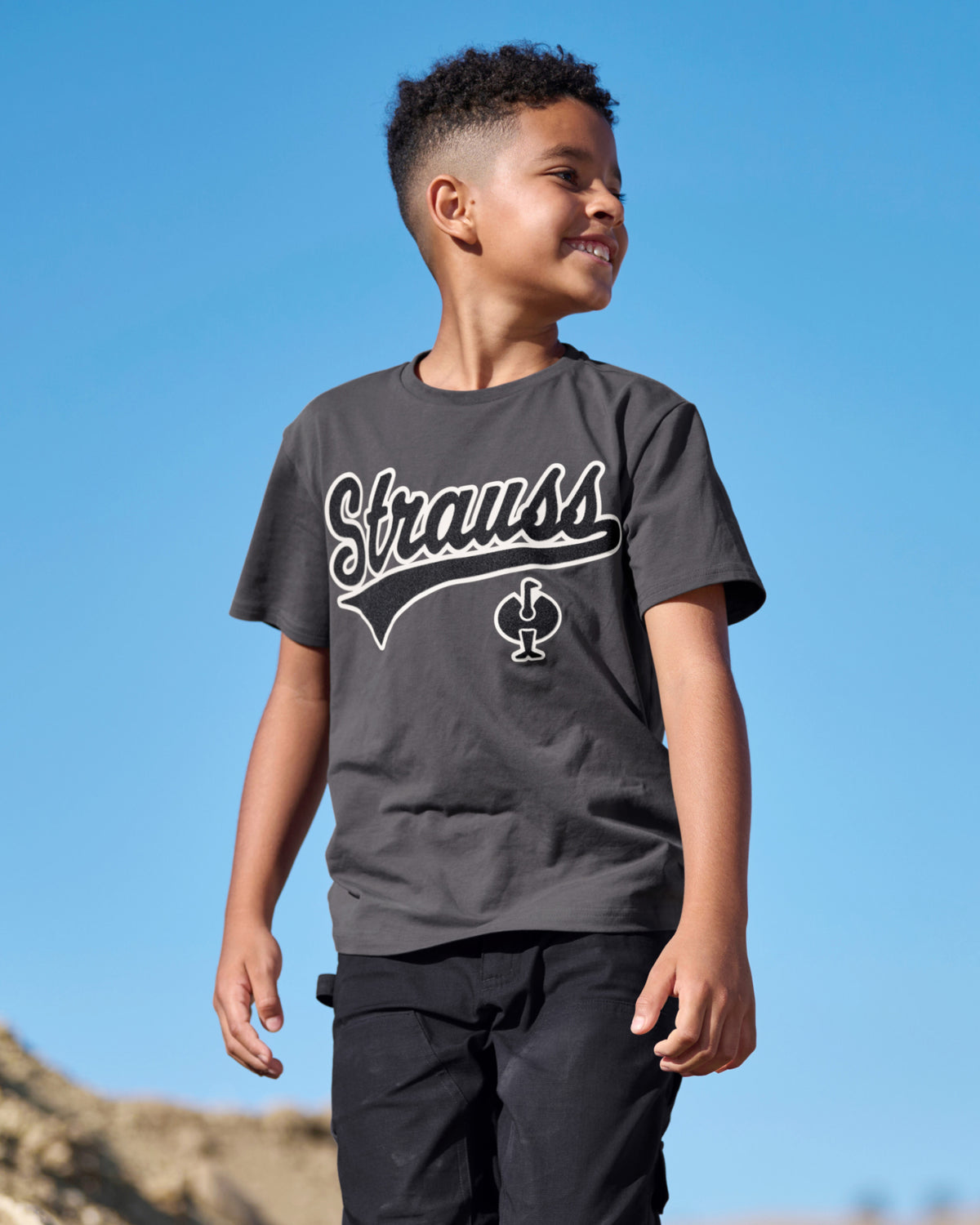 A child wears the Kids' STRAUSS Artwork T-Shirt e.s.e:pic in carbon gray under a clear blue sky