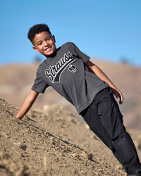A child wears the Kids' STRAUSS Artwork T-Shirt e.s.e:pic in carbon gray while holding a rock in a dry landscape