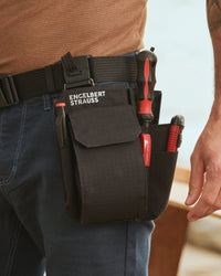 A black STRAUSS e.s.tool concept multipocket is fastened to work pants while a worker reaches for hand tools.