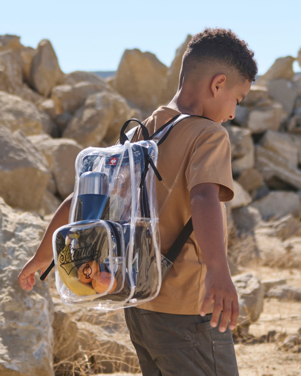 A child wears a clear STRAUSS e.s.e:pic backpack over a beige shirt with work pants.
