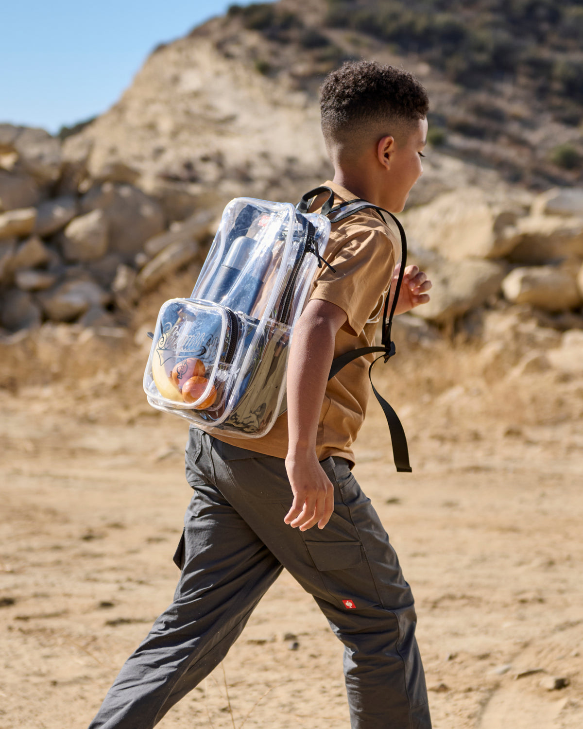 A person wears a clear STRAUSS e.s.e:pic backpack over a shirt and utility pants outdoors.