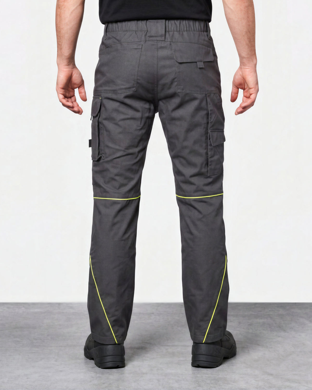 Rear view of a person wearing dark gray cargo work pants with bright yellow-green contrast stitching, paired with a black shirt and black shoes, against a light gray studio background.