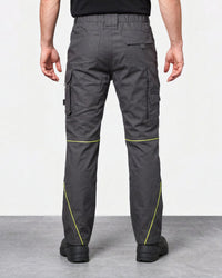 Rear view of a person wearing dark gray cargo work pants with bright yellow-green contrast stitching, paired with a black shirt and black shoes, against a light gray studio background.