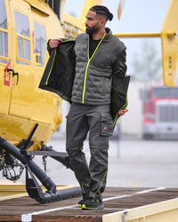 A man wearing a dark gray and black workwear jacket and matching cargo pants with neon yellow-green accents, standing on a wooden platform next to a yellow helicopter.