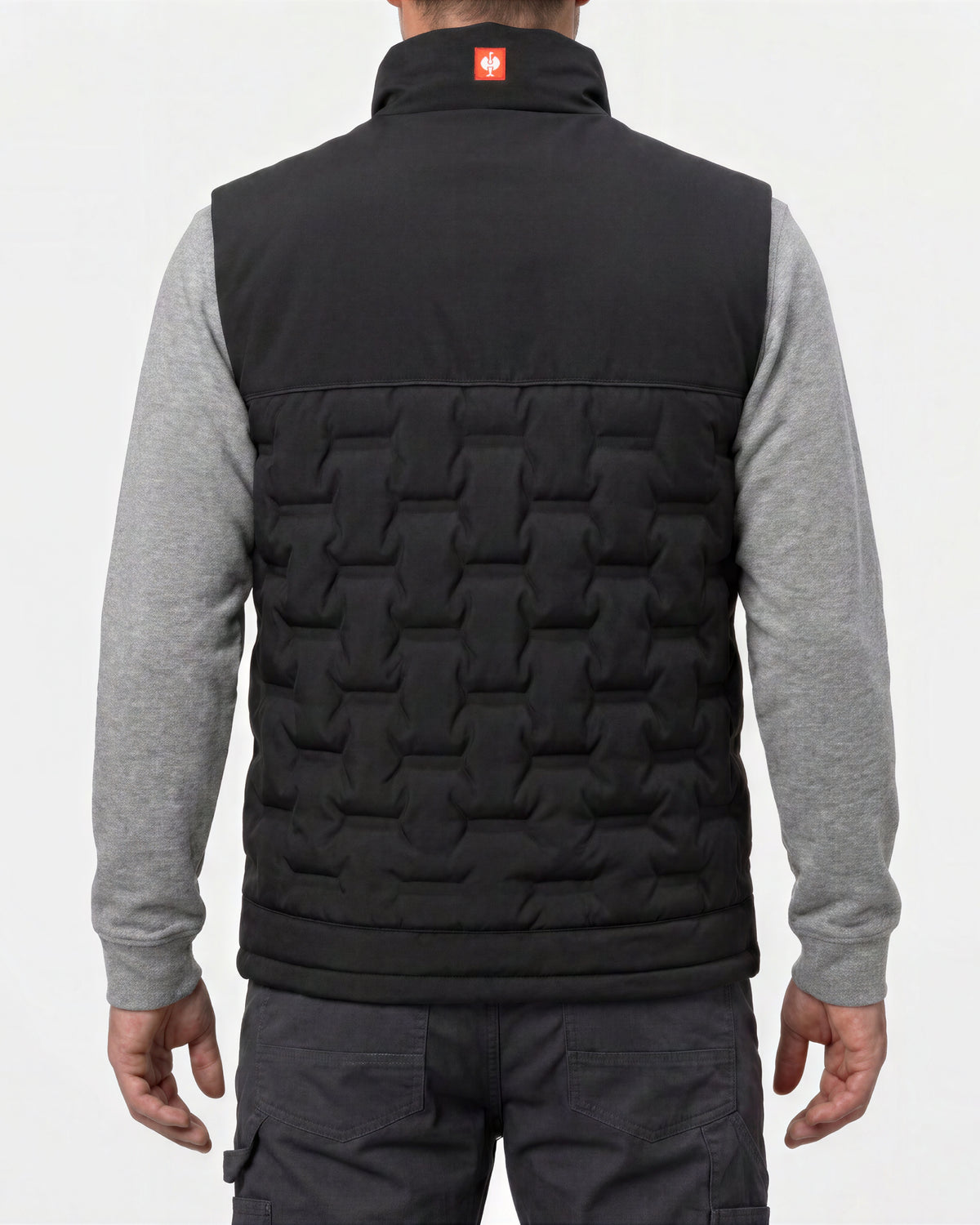 A man viewed from the back wearing a black quilted vest over a heather gray long-sleeve shirt and dark cargo pants, against a plain light gray studio background.