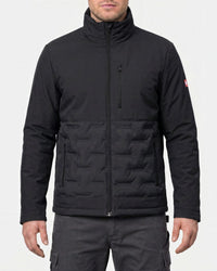 A man wearing a black technical jacket with a high collar, full zip front, and horizontal quilted paneling on the lower half, shown against a light gray studio background.
