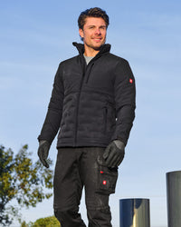 A man wearing a black technical fleece jacket, black work pants, and black gloves, standing outdoors against a blue sky.