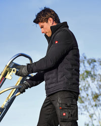 A man wearing a black insulated puffer jacket and black cargo pants with Swiss flag logo details, operating a metal cart outdoors against a blue sky.