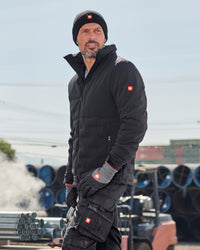 A man wearing a black technical jacket, black beanie, and black work gloves with a red Swiss cross logo, standing outdoors in front of a light blue sky and stacks of blue pipes.