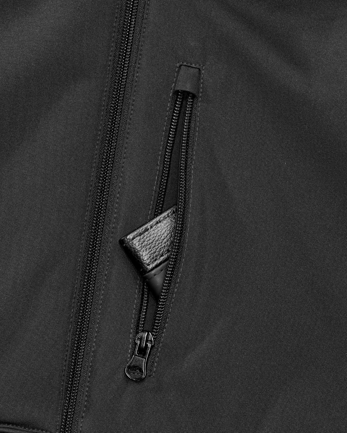 Close-up of a black zippered jacket or outerwear piece, showing a vertical front zipper and a diagonal zip pocket with a small black leather pull tab.