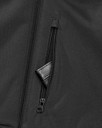 Close-up of a black zippered jacket or outerwear piece, showing a vertical front zipper and a diagonal zip pocket with a small black leather pull tab.