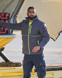 A man wearing a dark gray puffer jacket with bright neon green zippers and a matching dark cargo pants, standing next to a yellow aircraft on an airfield.
