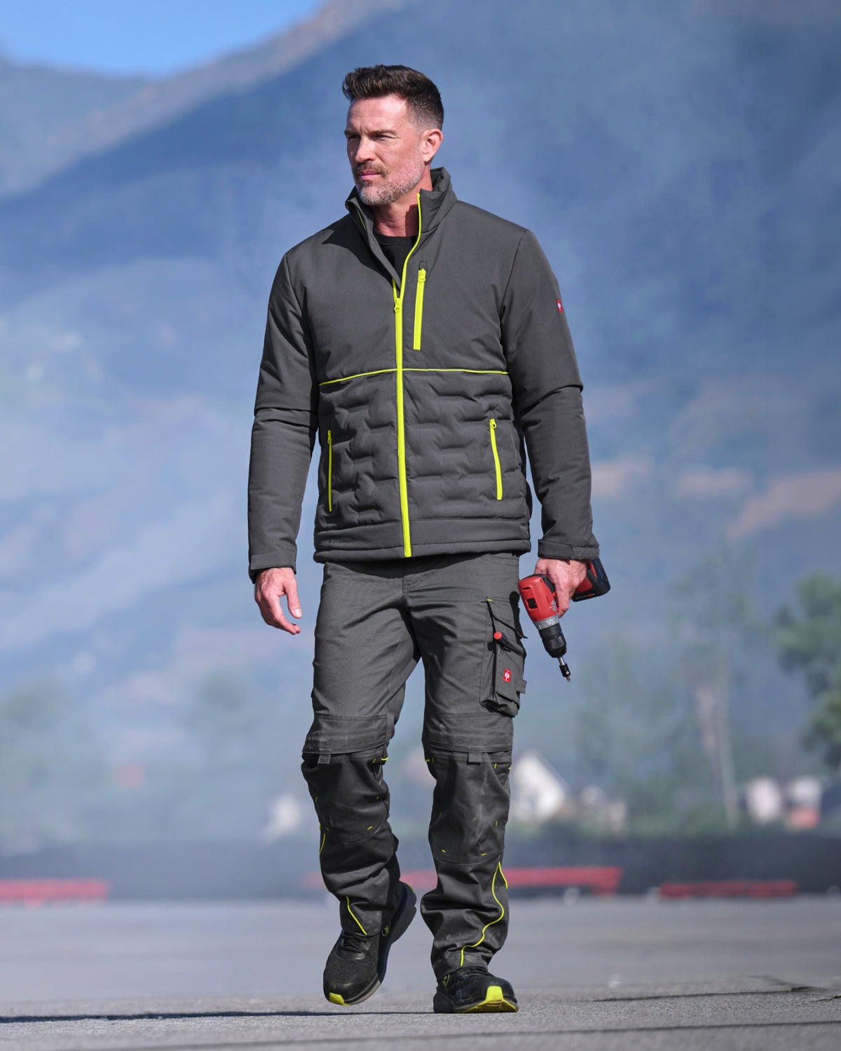 A man wearing a dark gray insulated jacket and matching dark gray work pants with neon yellow accents, walking outdoors on a paved surface with mountains in the background.