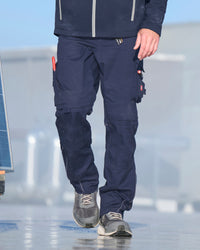 A person wearing dark navy blue work pants with multiple utility pockets and gray sneakers, shown from the waist down in an outdoor setting.