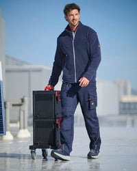 A man wearing a navy blue work jacket and matching cargo pants stands on a rooftop terrace, holding a black rolling tool case with red accents. The background shows a clear blue sky and distant industrial buildings.