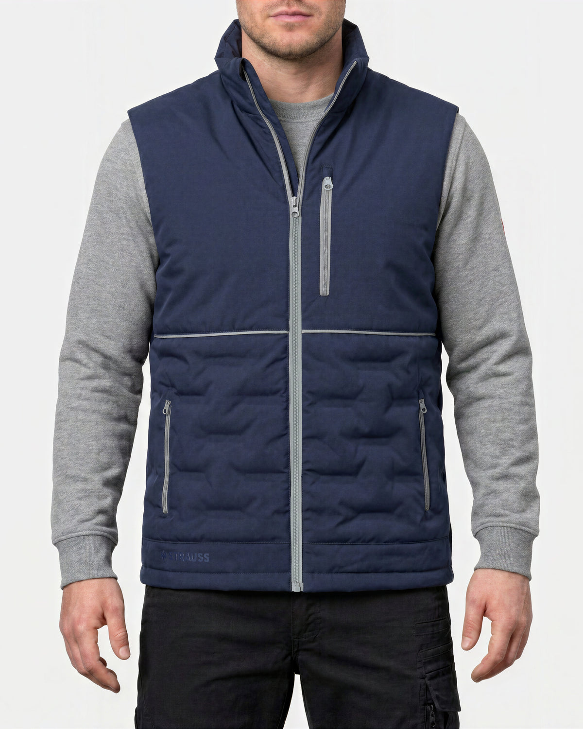 A man wearing a navy blue quilted vest with gray sleeves and gray zipper details, layered over a gray long-sleeve shirt and black pants, against a light gray background.