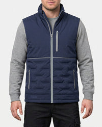 A man wearing a navy blue quilted vest with gray sleeves and gray zipper details, layered over a gray long-sleeve shirt and black pants, against a light gray background.