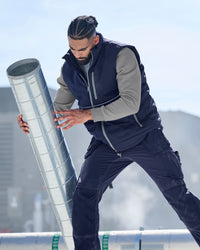 A man in a navy and gray work uniform pushes a large galvanized steel ventilation duct outdoors on a rooftop.