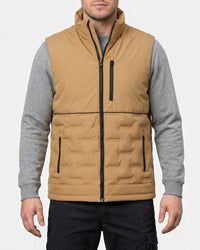 A man wearing a tan insulated zip-front vest over a gray long-sleeve shirt and black pants, shown from the lower face to upper thighs against a light gray studio background.