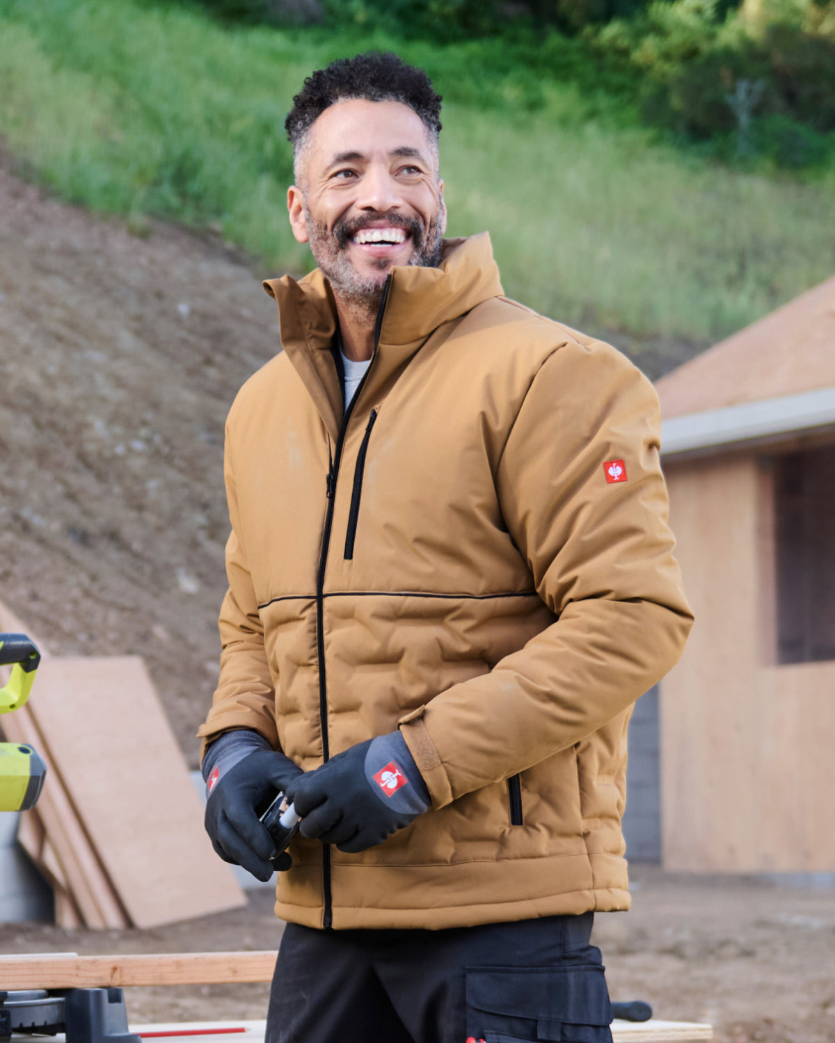 A smiling man wearing a tan insulated jacket with black gloves, standing outdoors in front of a wooden structure and grassy hillside.