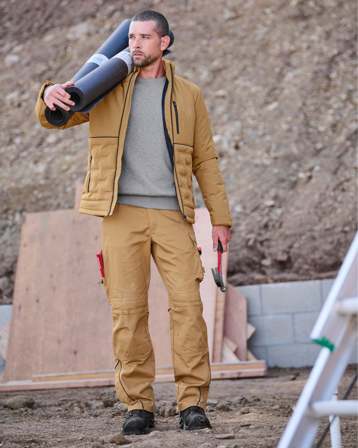 A man wearing a tan work jacket and matching work pants stands on a construction site, carrying a rolled-up mat over his shoulder and holding a hammer.