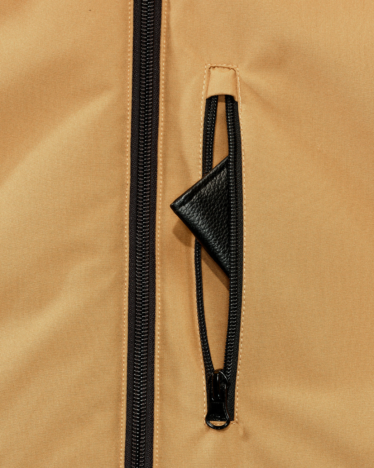 Close-up of a tan jacket with a black zipper and a black zippered pocket featuring a triangular black pull tab.