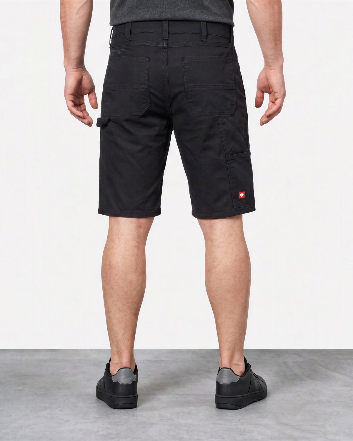 A man viewed from the back wearing black cargo shorts, a dark gray t-shirt, and black sneakers, standing against a white wall and gray floor.