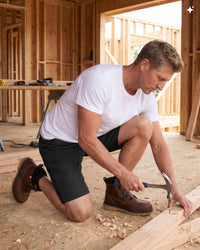 A man in a white t-shirt and black shorts kneels on a wood floor inside a construction site, using a hammer to secure a piece of wood. He is wearing brown work boots.