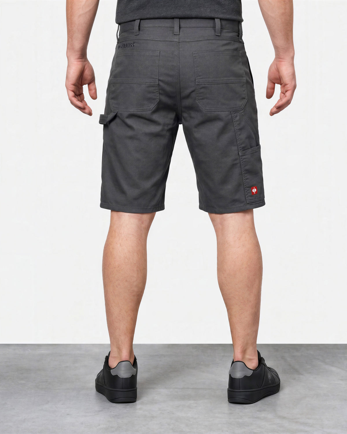 A man viewed from the back wearing dark gray cargo shorts, a dark gray t-shirt, and black slip-on shoes against a white studio background.