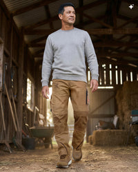 A man walking through a rustic barn wearing a light gray crewneck sweatshirt, tan work pants, and brown boots.