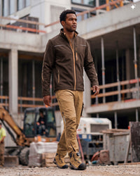 A man walking on a construction site wearing a brown zip-up work jacket, tan work pants, and tan work boots.