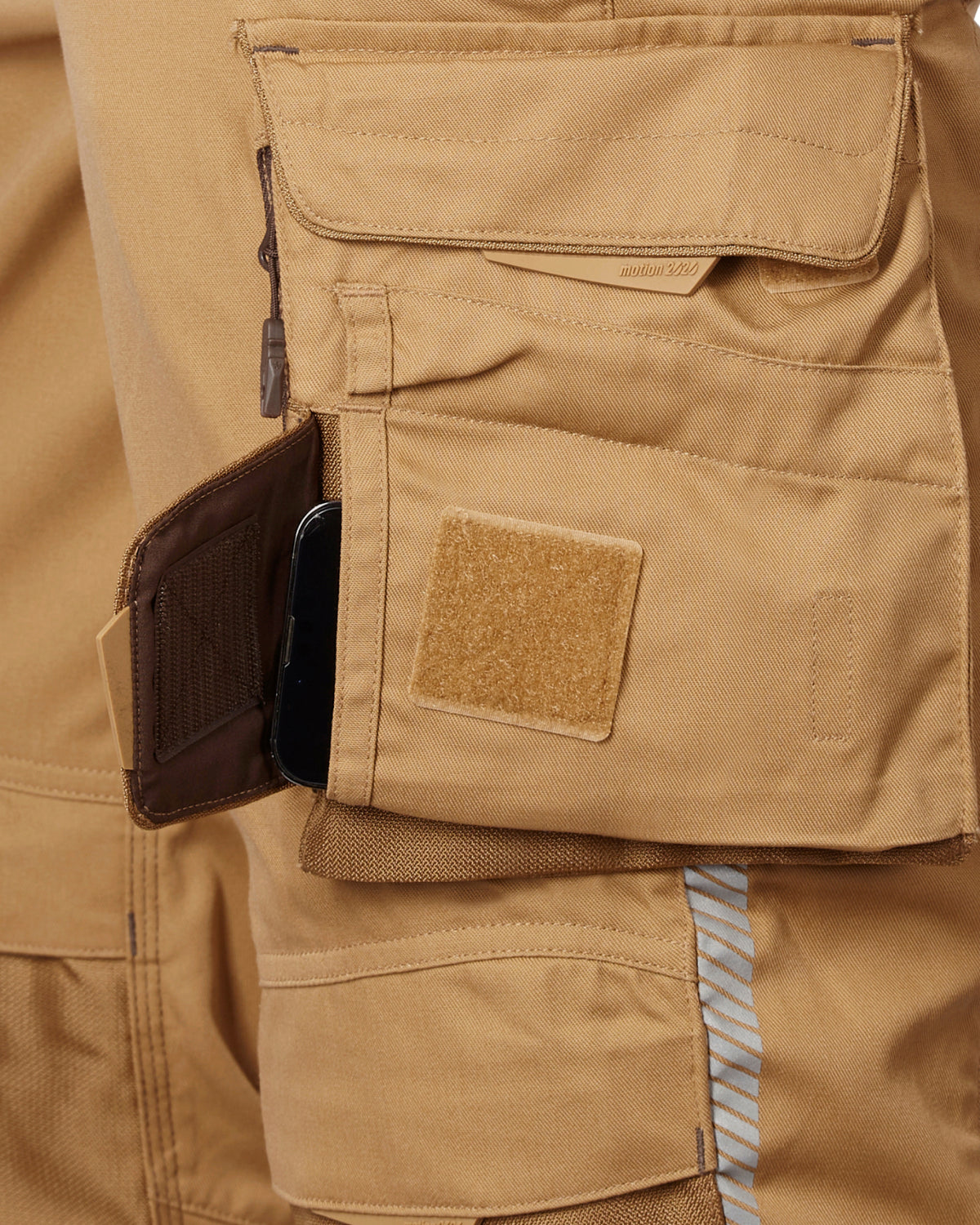 Close-up of a tan cargo pocket showing a side utility pocket containing a black smartphone, alongside a tan Velcro patch.