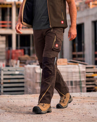 A man wearing dark brown and black technical work pants with utility pockets, a dark vest, and work boots, walking on a concrete surface at a construction site.