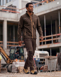 A man wearing a brown work jacket and matching work pants walks on a construction site, with construction equipment and a building structure blurred in the background.