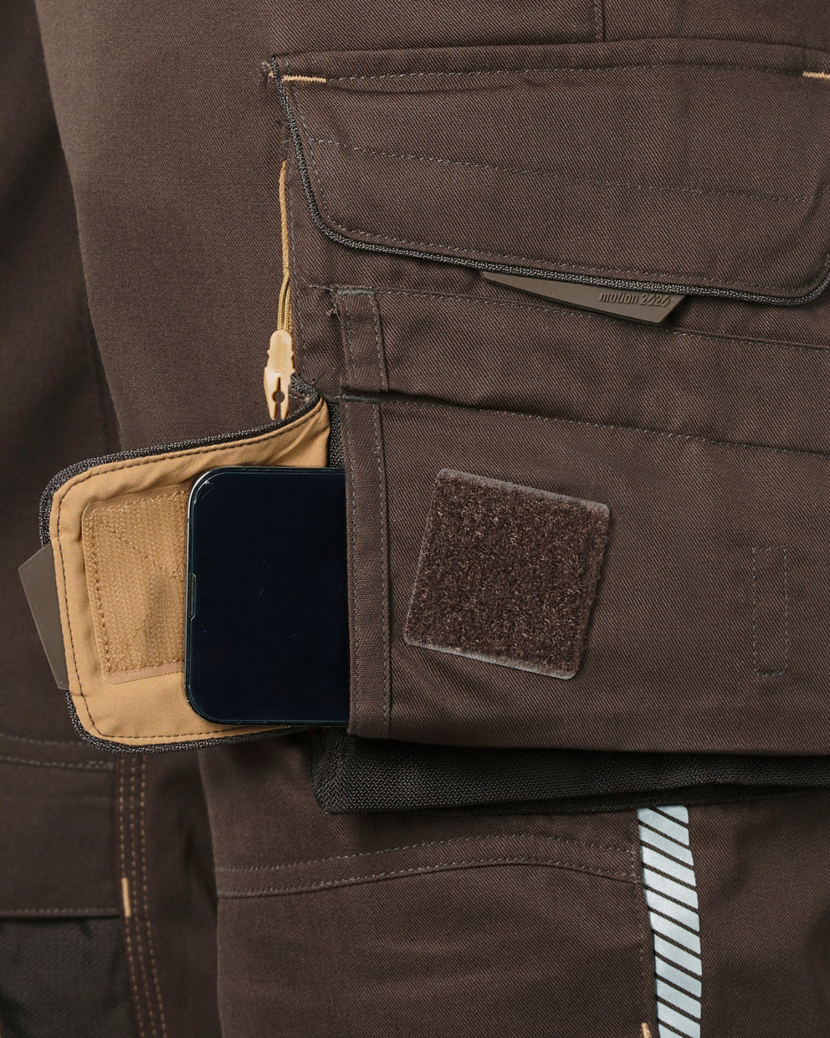 Close-up of a brown tactical utility pocket on a pants, showing a beige interior slip pocket with a black smartphone inserted and a brown hook-and-loop patch on the front.