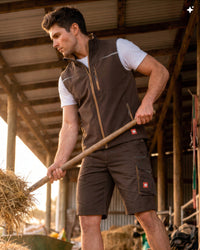 A man in a barn wearing a brown zip-up work vest and matching brown cargo shorts, holding a straw bale. The vest has a brown collar and zipper trim, and the shorts have brown zippers and side cargo pockets with red logo patches.