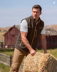 A man wearing a brown zip-front vest over a beige t-shirt and khaki pants, standing outdoors next to a hay bale with red barns in the background.