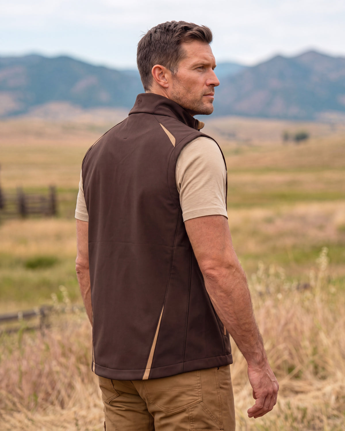 A man wearing a brown sleeveless zip-front vest over a beige short-sleeve shirt and khaki pants, standing in an outdoor field with mountains in the background.