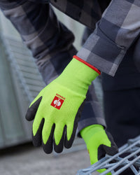 A worker wears neon green and black STRAUSS Senso-Grip latex knit gloves while gripping a hand tool at a workbench.