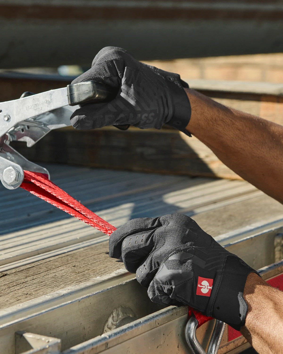 A worker wears black STRAUSS e.s.trail light graphic mechanic gloves while gripping a hand tool at a workbench.