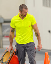 A worker wears the STRAUSS Enhanced Visibility Performance Polo Shirt in neon yellow with dark work pants near construction equipment