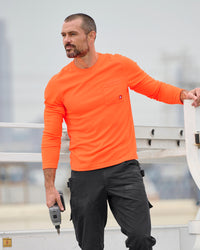 A worker wears the STRAUSS Enhanced Visibility Performance Long Sleeve Shirt in neon orange with dark work pants near construction equipment