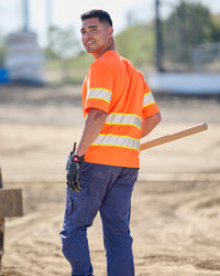 A worker wears the STRAUSS Class 2 High Visibility T-Shirt in neon orange with work pants while handling materials at a jobsite