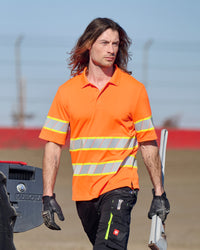 A worker wears the STRAUSS Class 2 High Visibility Polo Shirt in neon orange with dark work pants near construction equipment