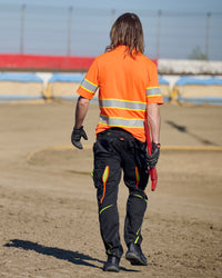 A worker wears the STRAUSS Class 2 High Visibility Polo Shirt in neon orange with work pants while handling materials at a jobsite