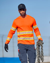 A worker wears the STRAUSS Class 2 High Visibility Long Sleeve Shirt in neon orange with dark work pants near construction equipment